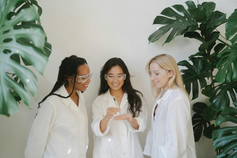 Three female scientists wearing goggles and white coats working together with plants in a lab.